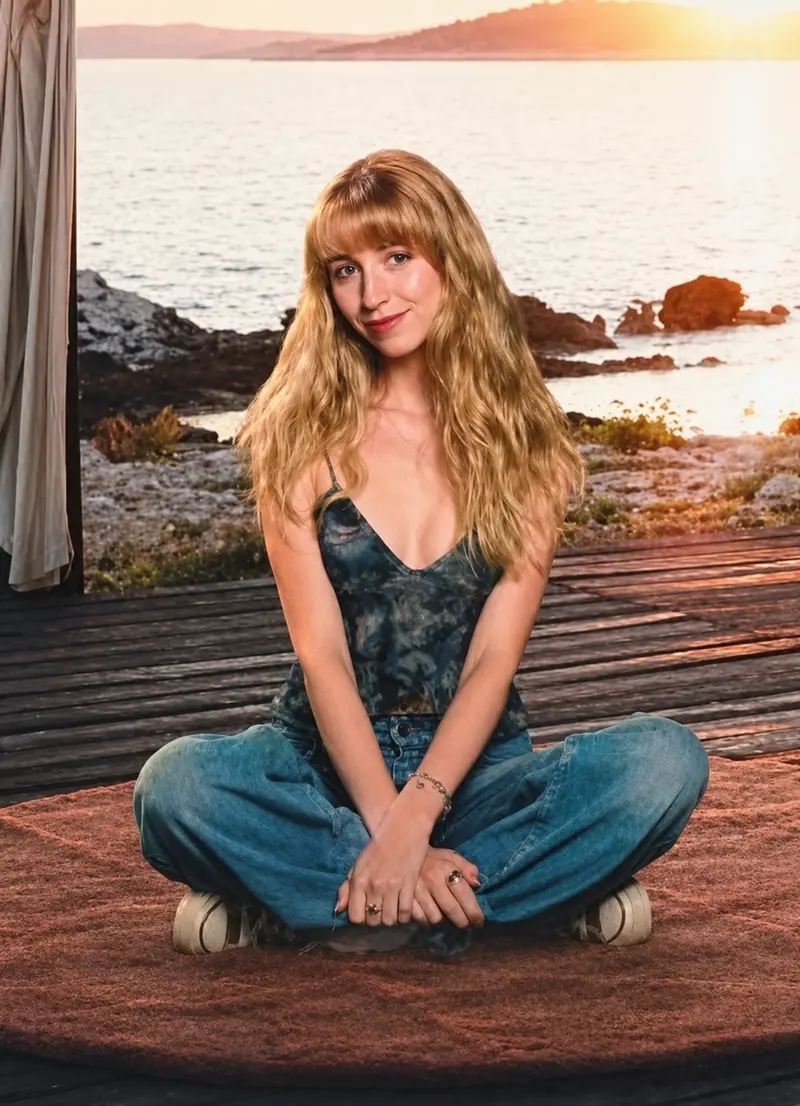 Joy sitting cross-legged on a wooden deck at sunset, smiling, with the sea behind her